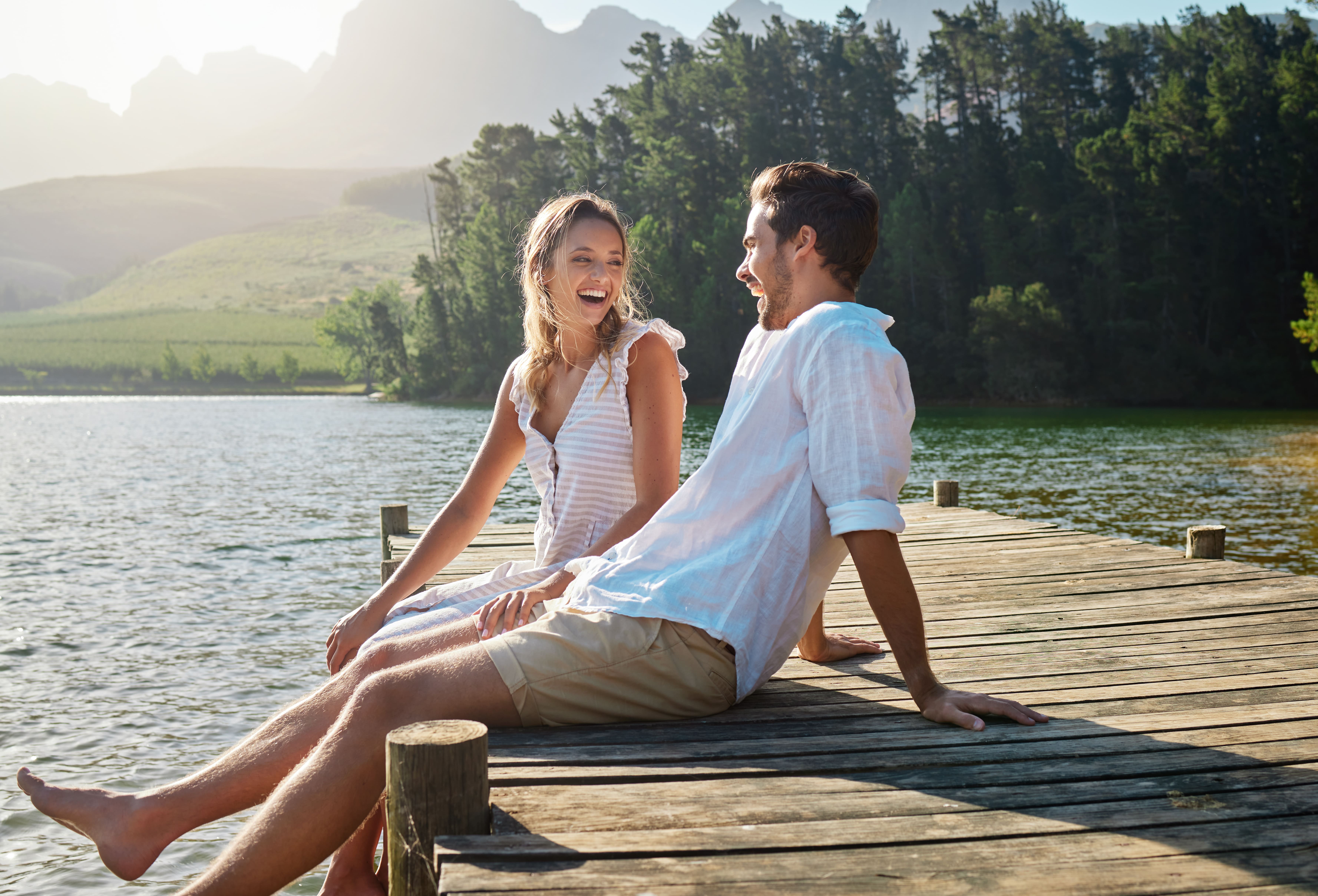casal sorrindo e se olhando sentados em pier de madeira ao redor de um lago com natureza ao fundo