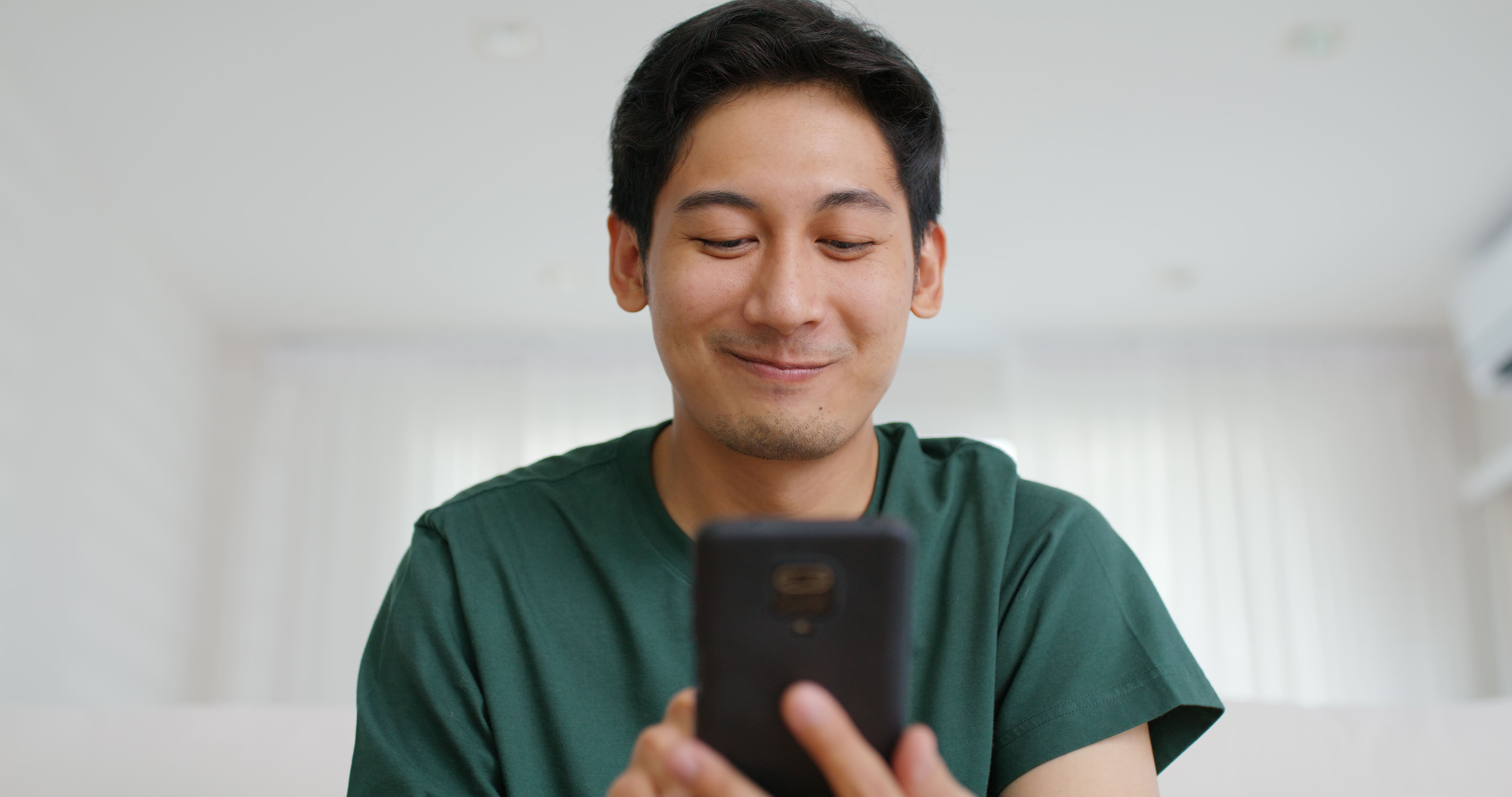 homem de camisa verde olhando a tela do celular enquanto dá um sorriso contido