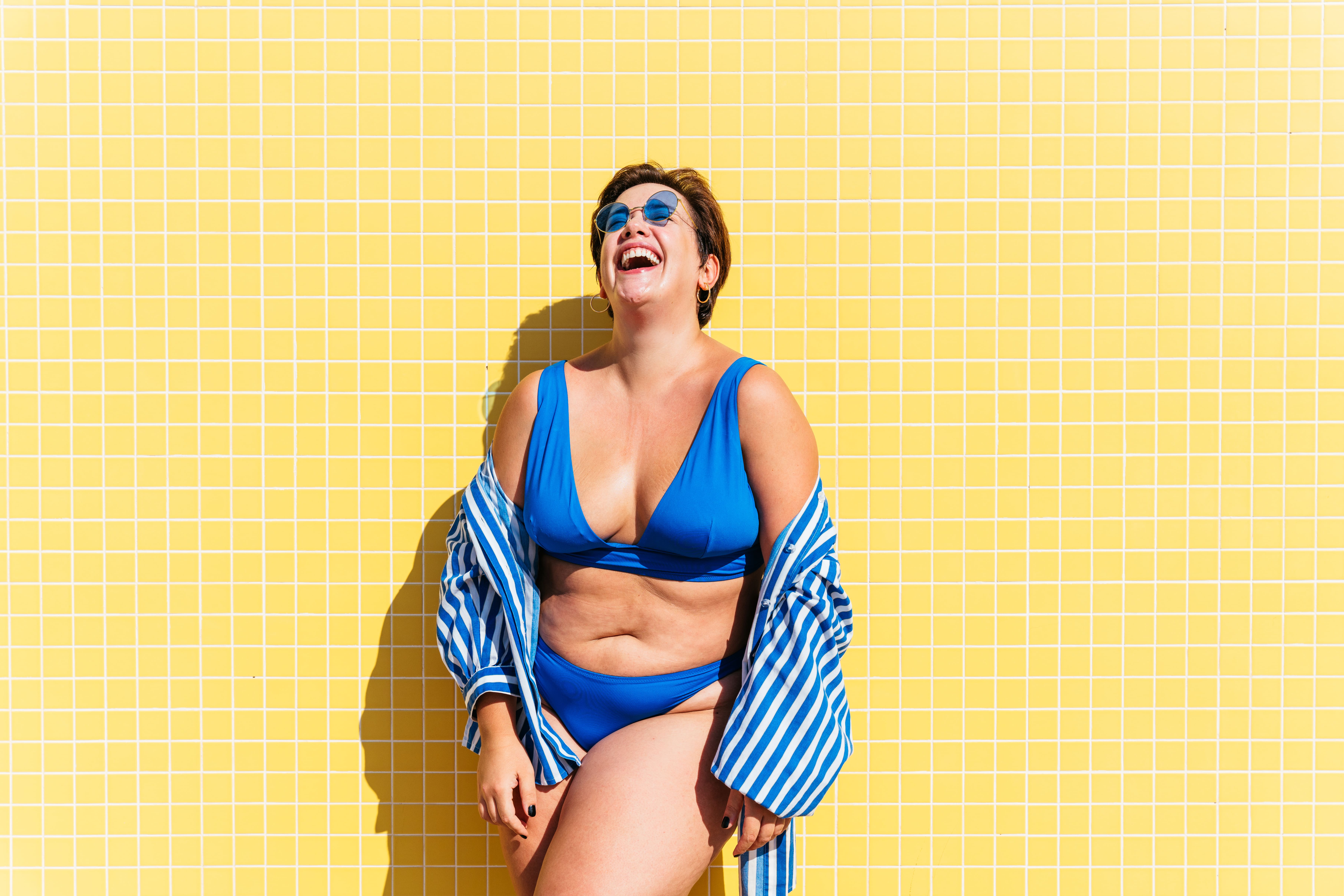 mulher bonita e confiante plus size sorrindo com roupas de praia azul e fundo amarelo 