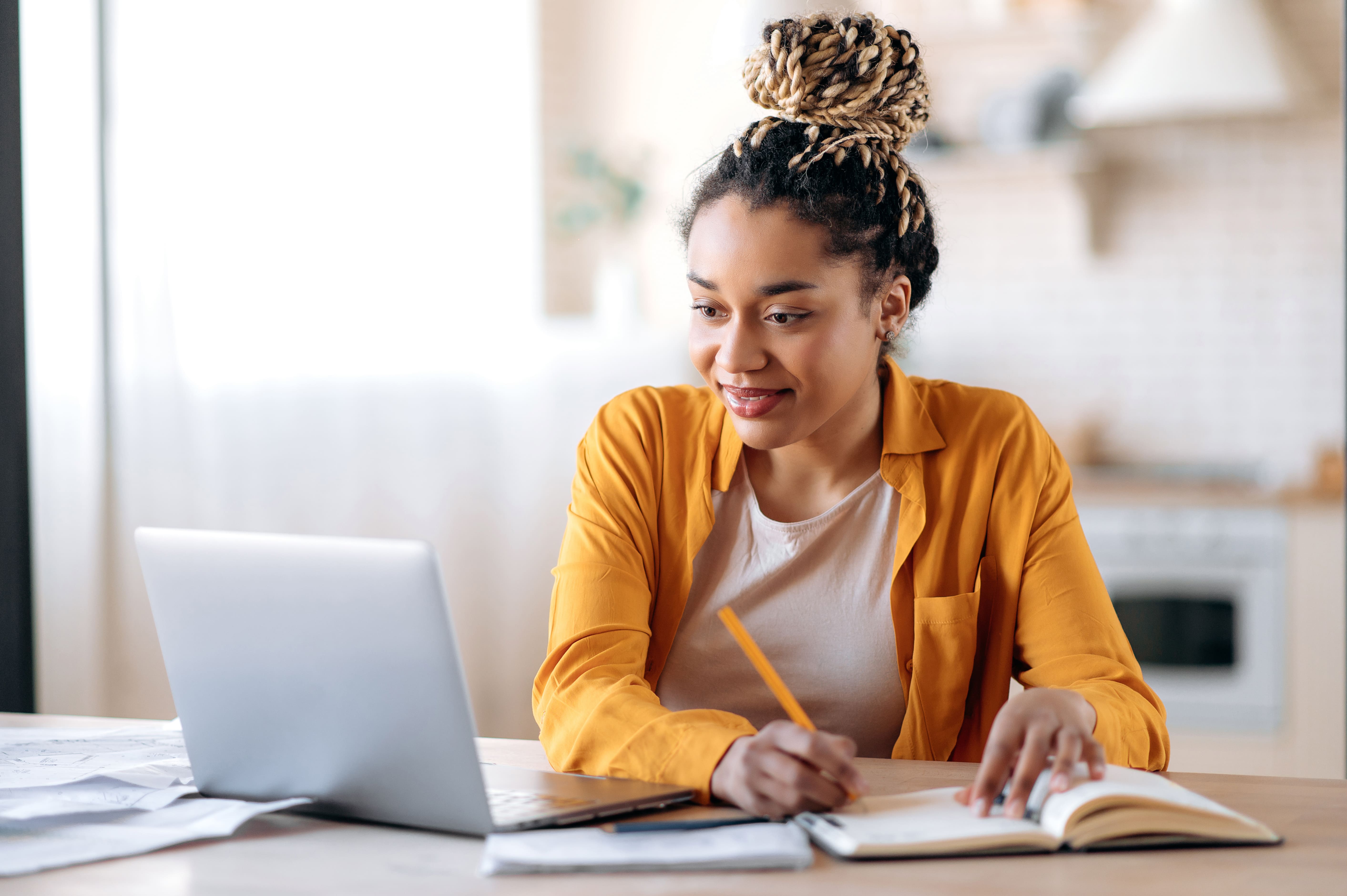 Mulher afroamericana com dreadlocks estudando remotamente no notebook em casa tomando notas enquanto está atenta e sorri