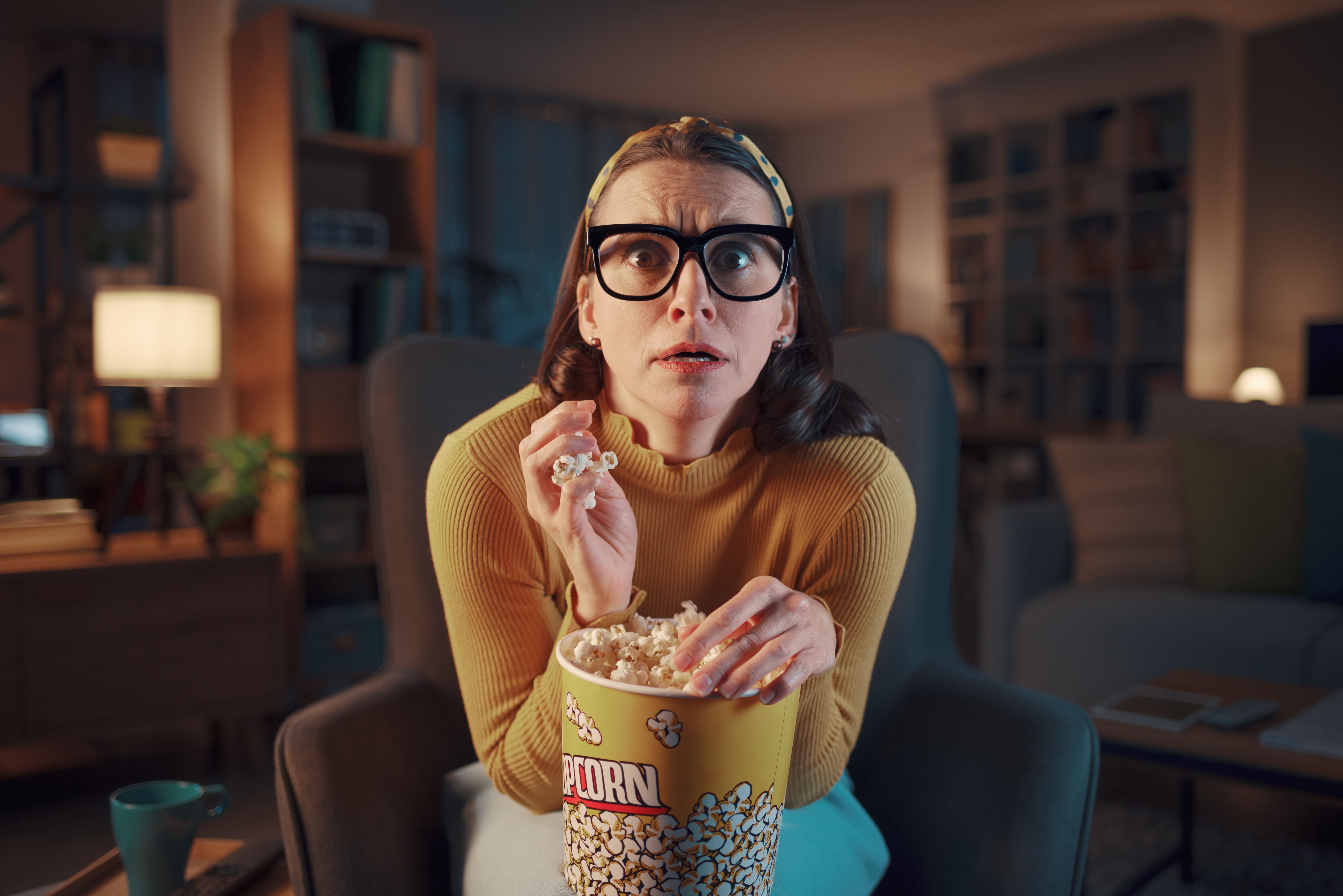 Mulher com expressão assustada comendo pipoca enquanto assiste televisão na sala.