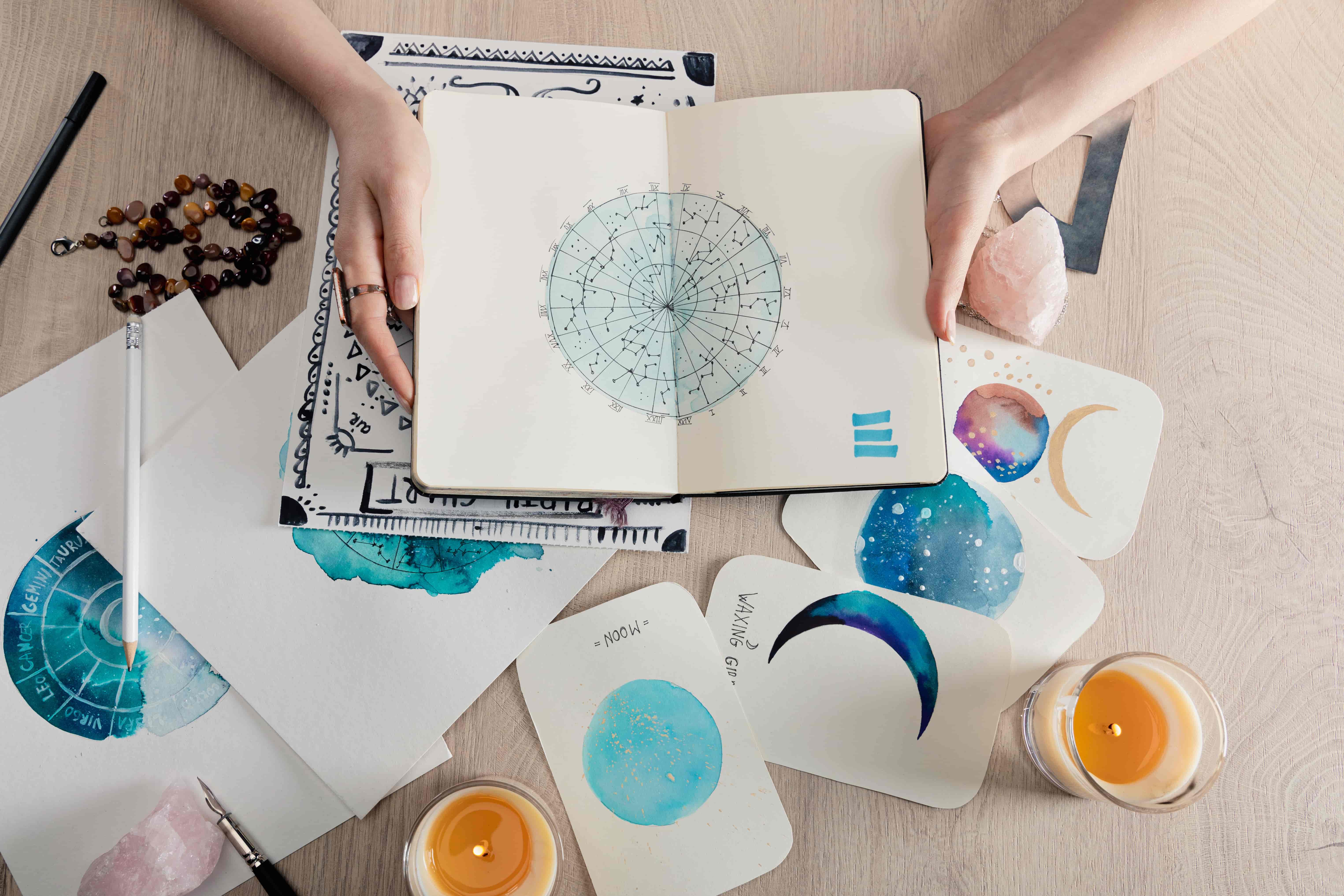 vista de cima de astrólogo segurando notebook com desenhos em aquarela e cartas de signos do zodíaco em cima da mesa